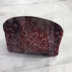 Puede incluir: Un neceser burdeos con estampado de cachemira y cremallera dorada. La bolsa tiene forma de media luna y presenta un patrón de pañuelo clásico en blanco. La bolsa está hecha de un material suave y acolchado.