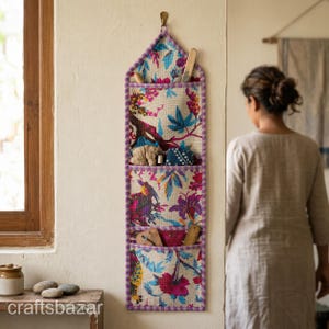 May include: A vertical wall-hanging fabric organiser featuring three tiered pockets. The textile has a cream background with a vibrant floral and bird pattern in shades of magenta, blue, and brown. The edges are finished with a purple trim.