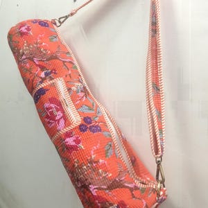 May include: An orange yoga mat bag with a floral pattern in pink, purple, and brown. The bag has a matching strap and white trim. The bag is hanging against a white wall.