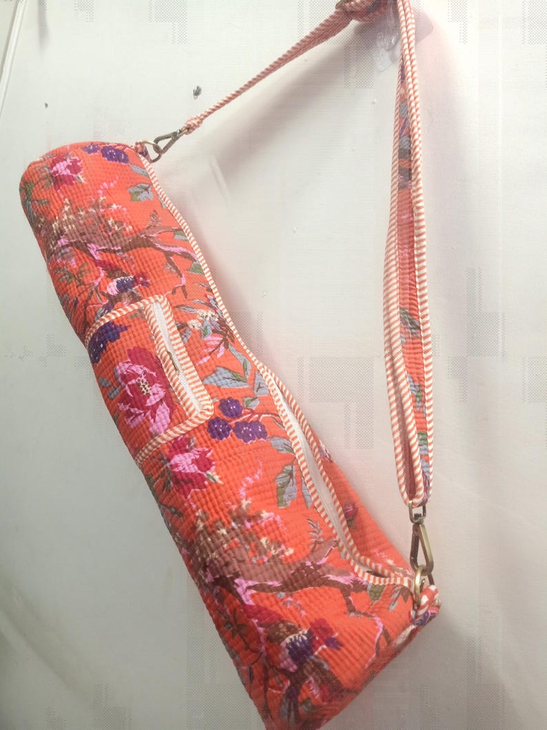 May include: An orange yoga mat bag with a floral pattern in pink, purple, and brown. The bag has a matching strap and white trim. The bag is hanging against a white wall.