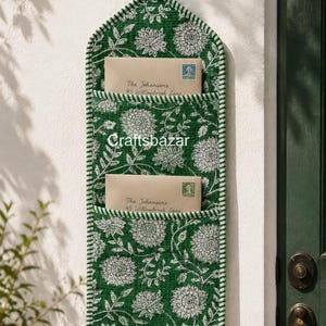 May include: A quilted fabric wall organiser featuring a green and white floral pattern with two pockets, each holding an envelope addressed to The Johansens at 45 Willowbrook Lane. The organiser has a striped green and white border and a hanging loop.