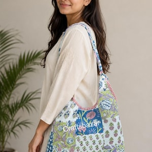 Könnte beinhalten: Eine gesteppte Hobo-Tasche mit Patchwork-Design, bestehend aus verschiedenen Blumen- und geometrischen Mustern in Blau-, Grün-, Rosa- und Weißtönen. Die Tasche hat einen breiten Schulterriemen und der Text Craftsbazar ist vorne aufgedruckt.