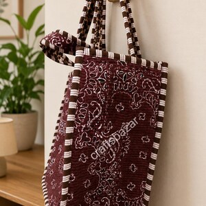 May include: A quilted tote bag in a deep maroon colour featuring a white paisley bandana pattern. The bag has brown and white striped fabric trim along the edges and handles. The text craftsbazar is printed in white on the side of the bag.