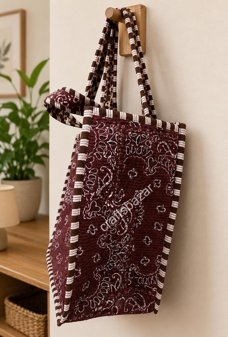 May include: A quilted tote bag in a deep maroon colour featuring a white paisley bandana pattern. The bag has brown and white striped fabric trim along the edges and handles. The text craftsbazar is printed in white on the side of the bag.