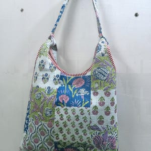 Könnte beinhalten: Eine Patchwork-Tragetasche mit einem Blumenmuster in Blau-, Grün- und Rosatönen. Die Tasche hat eine rote Bordüre und einen passenden Riemen. Die Tasche hängt an einer weißen Wand.