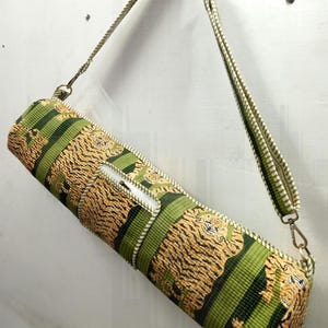 May include: A cylindrical yoga mat bag with a tiger stripe pattern in shades of tan and black, accented with green stripes. The bag has a white zipper and a matching shoulder strap with metal clasps.
