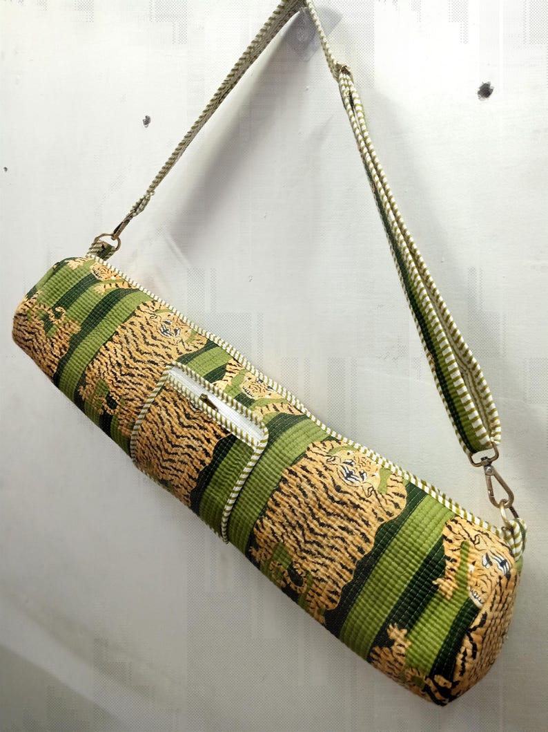 May include: A cylindrical yoga mat bag with a tiger stripe pattern in shades of tan and black, accented with green stripes. The bag has a white zipper and a matching shoulder strap with metal clasps.