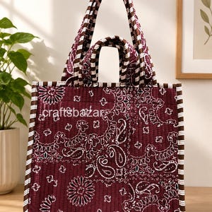 May include: A quilted tote bag in deep maroon featuring a white paisley pattern and brown and white striped trim. The bag has two sets of handles and the text craftsbazar is printed in white on the front. The fabric has a vertical stitched texture.