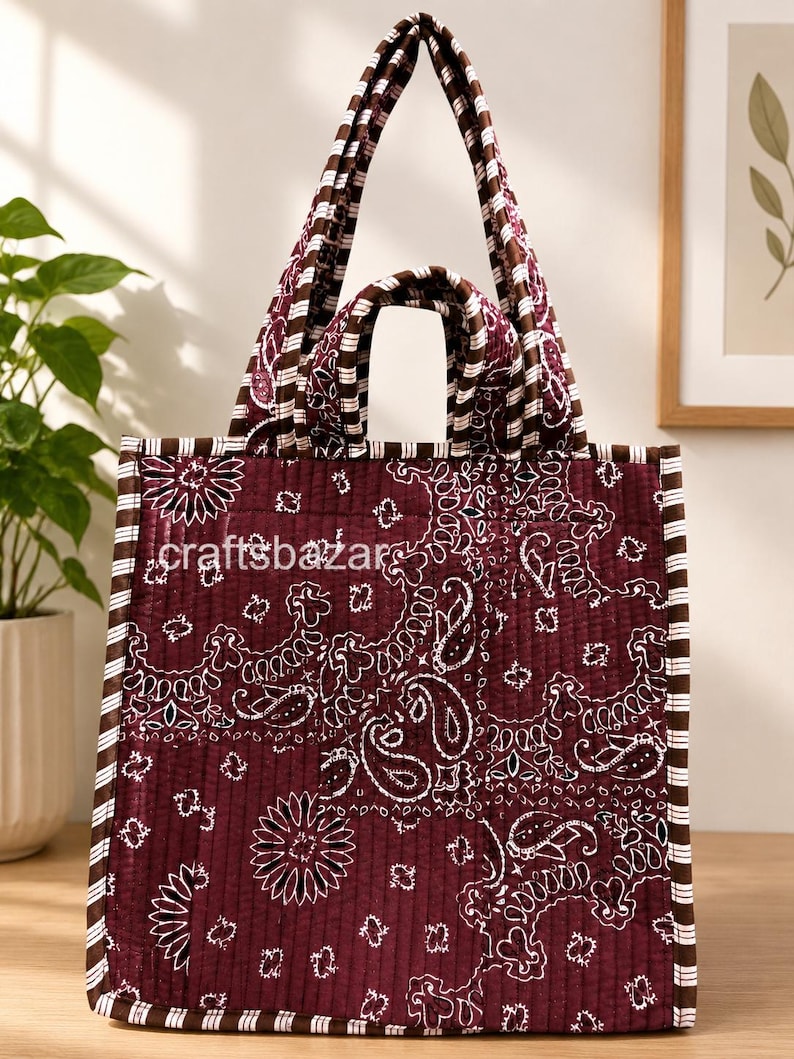 May include: A quilted tote bag in deep maroon featuring a white paisley pattern and brown and white striped trim. The bag has two sets of handles and the text craftsbazar is printed in white on the front. The fabric has a vertical stitched texture.