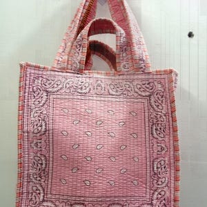 Könnte beinhalten: Eine rosa Tragetasche mit Paisley-Muster und gestreiftem Rand. Die Tasche hat zwei Griffe und ist aus gestepptem Stoff gefertigt. Der Hauptteil der Tasche zeigt ein großes Paisley-Design in quadratischer Form.