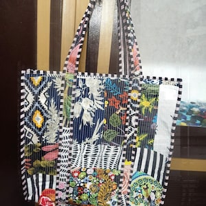 Könnte beinhalten: Eine Patchwork-Tragetasche mit einer Vielzahl farbenfroher Stoffmuster. Die Tasche weist eine Mischung aus floralen, geometrischen und gestreiften Designs in Blau-, Rot-, Grün- und Weißtönen auf. Die Griffe und Kanten sind mit schwarz-weißen Streifen versehen.