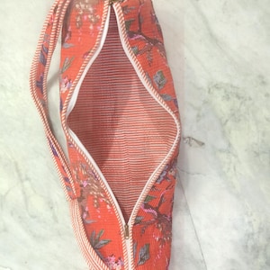 May include: An open, cylindrical yoga mat bag with a floral pattern in shades of orange, pink, and purple. The bag has a white zipper, a striped strap, and a white interior with red stripes. The bag is designed to carry a yoga mat.
