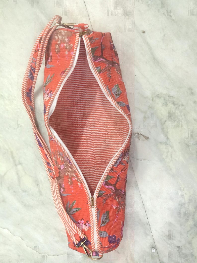 May include: An open, cylindrical yoga mat bag with a floral pattern in shades of orange, pink, and purple. The bag has a white zipper, a striped strap, and a white interior with red stripes. The bag is designed to carry a yoga mat.