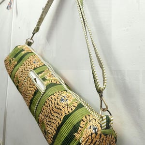 May include: A cylindrical yoga mat bag with a tiger stripe pattern in shades of green and tan. The bag has a zipper closure and a matching adjustable shoulder strap with metal clasps. The bag is hanging against a white wall.