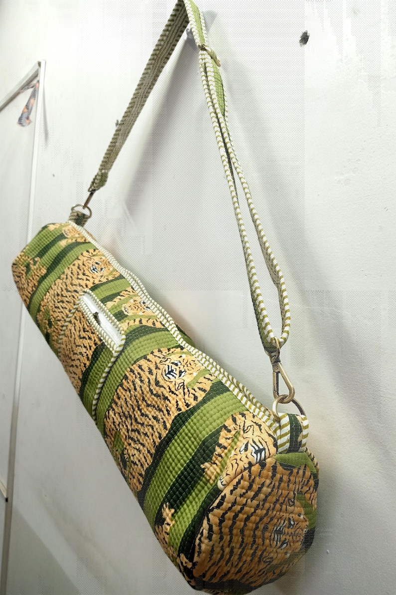May include: A cylindrical yoga mat bag with a tiger stripe pattern in shades of green and tan. The bag has a zipper closure and a matching adjustable shoulder strap with metal clasps. The bag is hanging against a white wall.