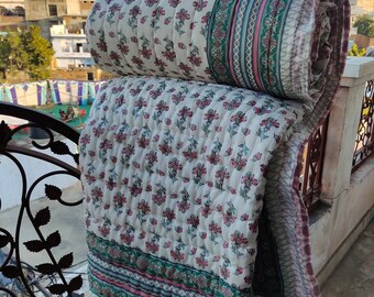May include: A rolled-up, floral-patterned quilt with a white background and pink flowers. The quilt has a green and pink border and is displayed outdoors. The quilt is made of a soft, quilted material.