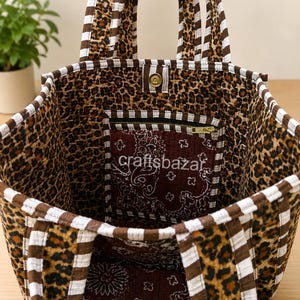 May include: A fabric tote bag featuring a brown and black leopard print exterior with white and brown striped trim and handles. The interior is lined with a dark maroon paisley fabric and includes a zippered pocket with the text craftsbazar.