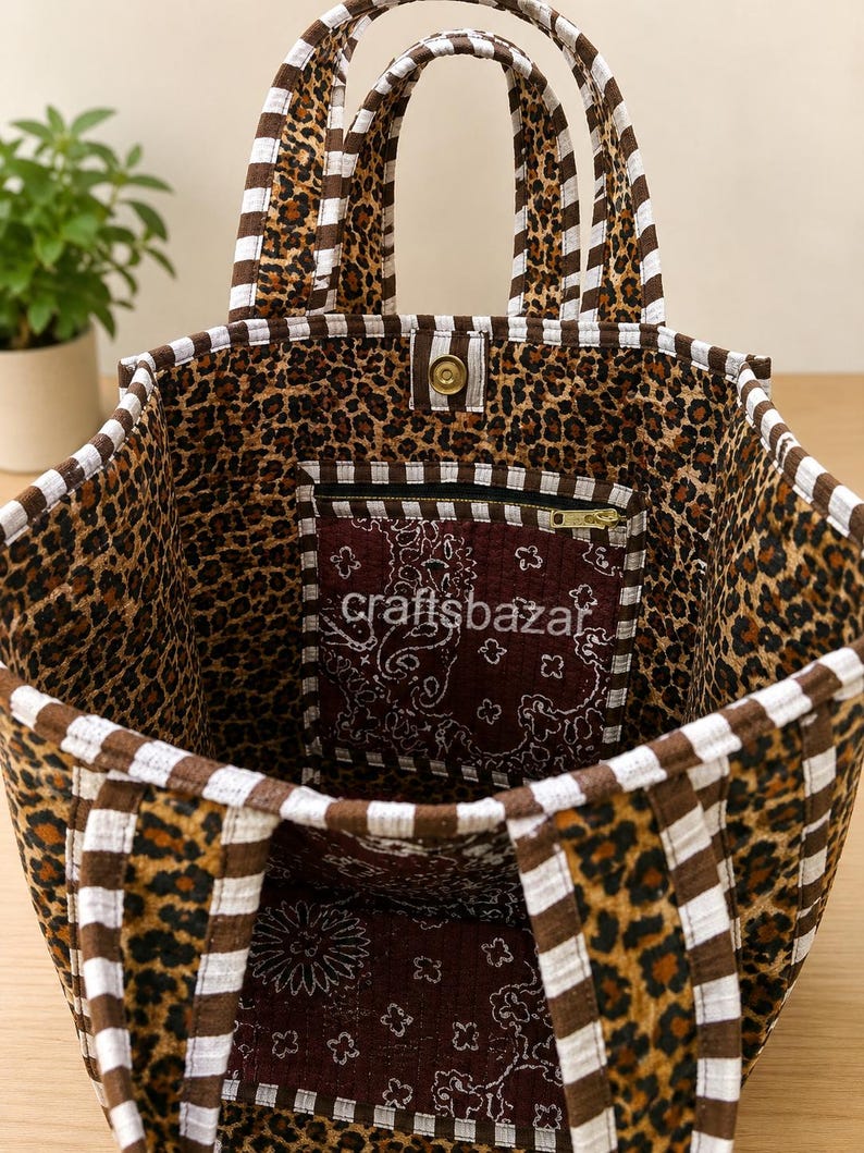 May include: A fabric tote bag featuring a brown and black leopard print exterior with white and brown striped trim and handles. The interior is lined with a dark maroon paisley fabric and includes a zippered pocket with the text craftsbazar.