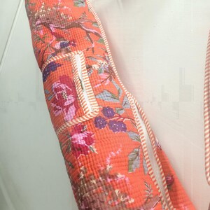 May include: An orange yoga mat bag with a floral pattern featuring pink, purple, and brown flowers and branches. The bag has a white zipper and trim with orange stripes. A strap is attached for carrying.