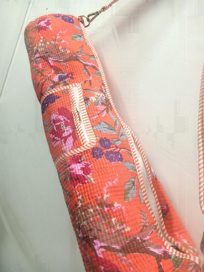 May include: An orange yoga mat bag with a floral pattern featuring pink, purple, and brown flowers and branches. The bag has a white zipper and trim with orange stripes. A strap is attached for carrying.