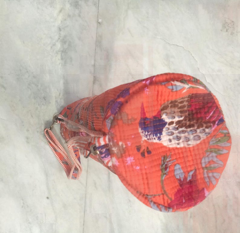 May include: An orange cylindrical bag with a bird and floral pattern. The bag has a zipper closure and a small handle. The bird is depicted with blue, brown, and red colors. The floral pattern includes red and purple flowers.