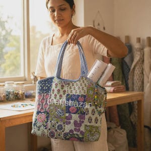 Könnte beinhalten: Eine gesteppte Tragetasche mit einem Patchwork-Design aus verschiedenen Blumen- und geometrischen Mustern in Blau-, Grün-, Rosa- und Violetttönen. Die Tasche hat zwei blau-weiß gestreifte Griffe und den Schriftzug craftsbazar auf der Vorderseite.