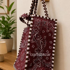 May include: A quilted tote bag in deep maroon with a white paisley pattern, featuring brown and white striped trim along the edges and handles. The text craftsbazar is printed in white on the side of the bag, which hangs from a wooden wall hook.