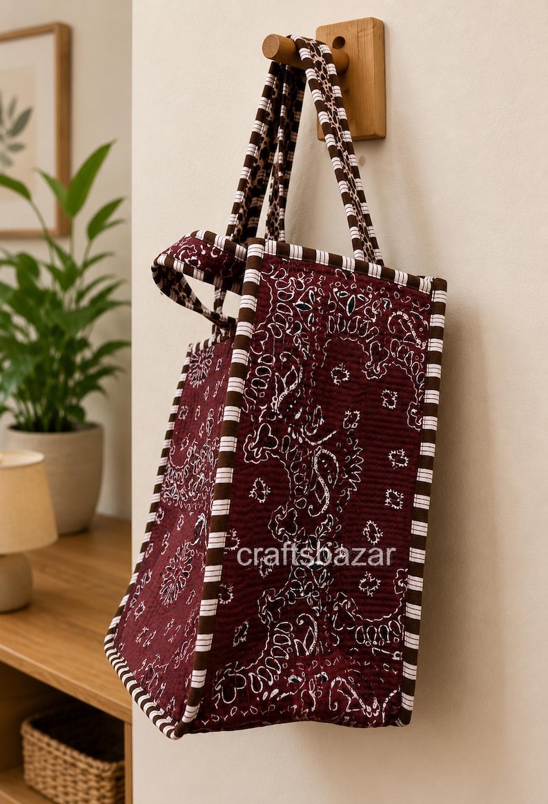 May include: A quilted tote bag in deep maroon with a white paisley pattern, featuring brown and white striped trim along the edges and handles. The text craftsbazar is printed in white on the side of the bag, which hangs from a wooden wall hook.