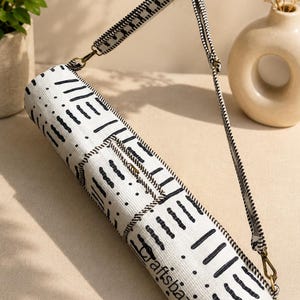 May include: A cylindrical yoga mat bag in white fabric with a black dashed pattern and the text "Craftsbazar" printed on the side. The bag features a long, adjustable black and white striped shoulder strap with brass-toned metal hardware clips.