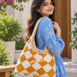 Könnte beinhalten: Eine Hobo-Schultertasche mit gestepptem Rautenmuster in Senfgelb und Off-White. Die Tasche verfügt über einen breiten, integrierten Trageriemen und einen kleinen metallischen Knopfverschluss in der Mitte. Der Text Craftsbazar ist vorne aufgedruckt.