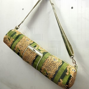 May include: A cylindrical yoga mat bag with a green and tan tiger stripe pattern. The bag has a zipper closure and a matching green and tan shoulder strap. The bag is hanging on a white wall.