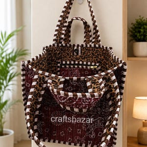 May include: A quilted tote bag featuring a mix of brown and white striped trim, a dark red paisley pattern, and a leopard print interior. The bag has two top handles and a smaller front pocket with the text craftsbazar printed in white.