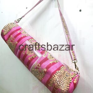 May include: A cylindrical yoga mat bag with a pink and gold tiger stripe pattern. The bag has a matching shoulder strap and metal hardware. The text "craftsbazar" is printed on the bag.