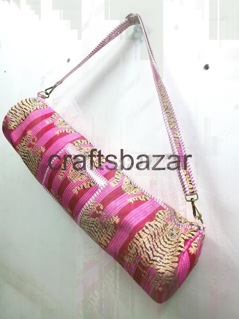 May include: A cylindrical yoga mat bag with a pink and gold tiger stripe pattern. The bag has a matching shoulder strap and metal hardware. The text "craftsbazar" is printed on the bag.