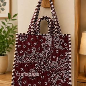 May include: A quilted tote bag in deep maroon featuring a white paisley bandana print. The bag has two sets of handles, a square silhouette, and brown and white striped trim along the edges. The text craftsbazar is printed on the front.