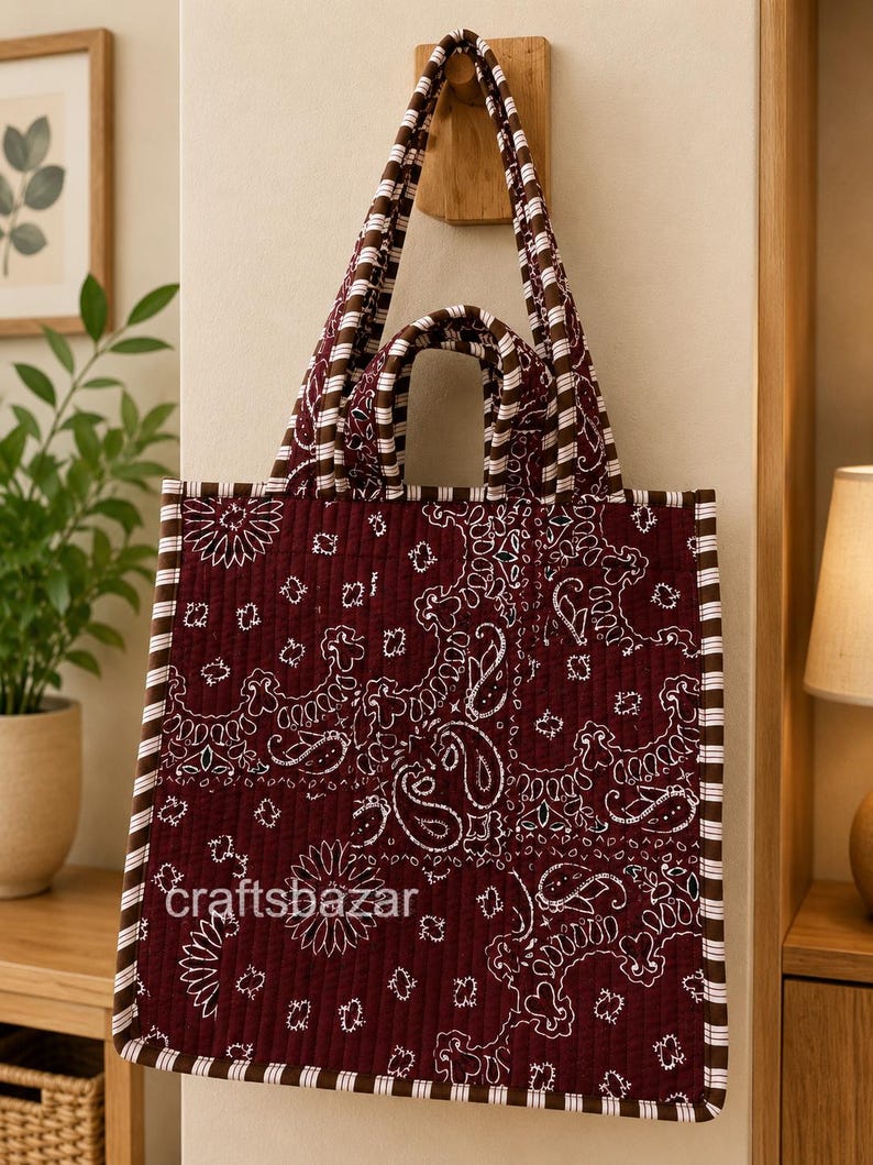 May include: A quilted tote bag in deep maroon featuring a white paisley bandana print. The bag has two sets of handles, a square silhouette, and brown and white striped trim along the edges. The text craftsbazar is printed on the front.