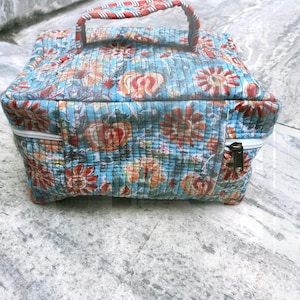 May include: A quilted, square-shaped bag with a floral pattern in shades of blue, red, and orange. The bag features a handle with red and white stripes and a zipper closure. The bag is designed for carrying items.