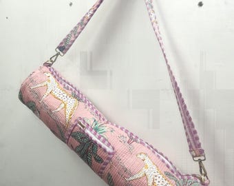 May include: A pink yoga mat bag with a repeating pattern of white cheetahs and tropical foliage. The bag has a zipper closure and a matching shoulder strap with metal hardware.