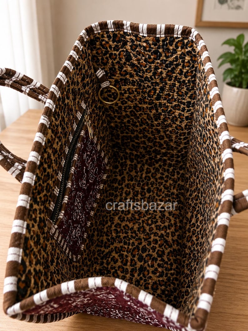 May include: A tote bag with a brown and white striped exterior trim and a leopard print interior lining. The bag features a dark maroon zippered pocket with a white floral pattern and a gold-toned metal ring. The text craftsbazar is visible.