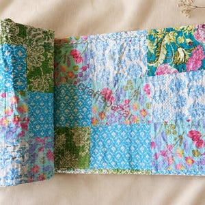Könnte beinhalten: Ein rechteckiges, gestepptes Textil mit einem Patchwork-Design aus verschiedenen Blumen- und geometrischen Mustern in Blau-, Grün- und Rosatönen. Der Stoff weist sichtbare Nähte auf und enthält in der Mitte den Schriftzug Craftsbazar.