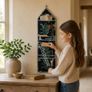 May include: A vertical wall-mounted fabric organizer featuring a black background with a green tropical leaf pattern and white striped trim. It includes multiple pockets for storage, with the word craftsbazar printed vertically in white text.