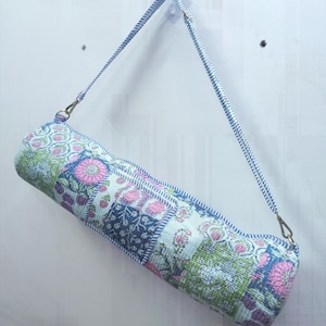 Könnte beinhalten: Eine zylindrische Yogamatten-Tasche mit einem Patchwork-Blumenmuster. Die Tasche hat rosa, grüne und blaue Blumenmuster und einen blau-weiß gestreiften Riemen. Die Tasche hängt an einer weißen Wand.