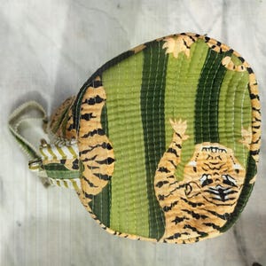 May include: A round, quilted bag with a tiger print. The bag features a green and yellow striped pattern with a tiger illustration. The bag has a white and green striped handle and a zipper closure.