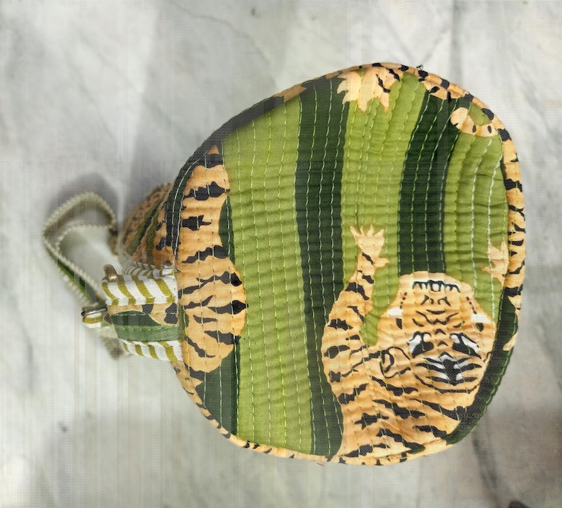 May include: A round, quilted bag with a tiger print. The bag features a green and yellow striped pattern with a tiger illustration. The bag has a white and green striped handle and a zipper closure.