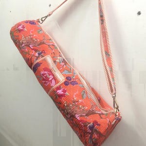 May include: An orange yoga mat bag with a floral pattern in pink, purple, and brown. The bag has a zipper closure and a matching strap with a rope-like design. The bag is hanging against a white wall.