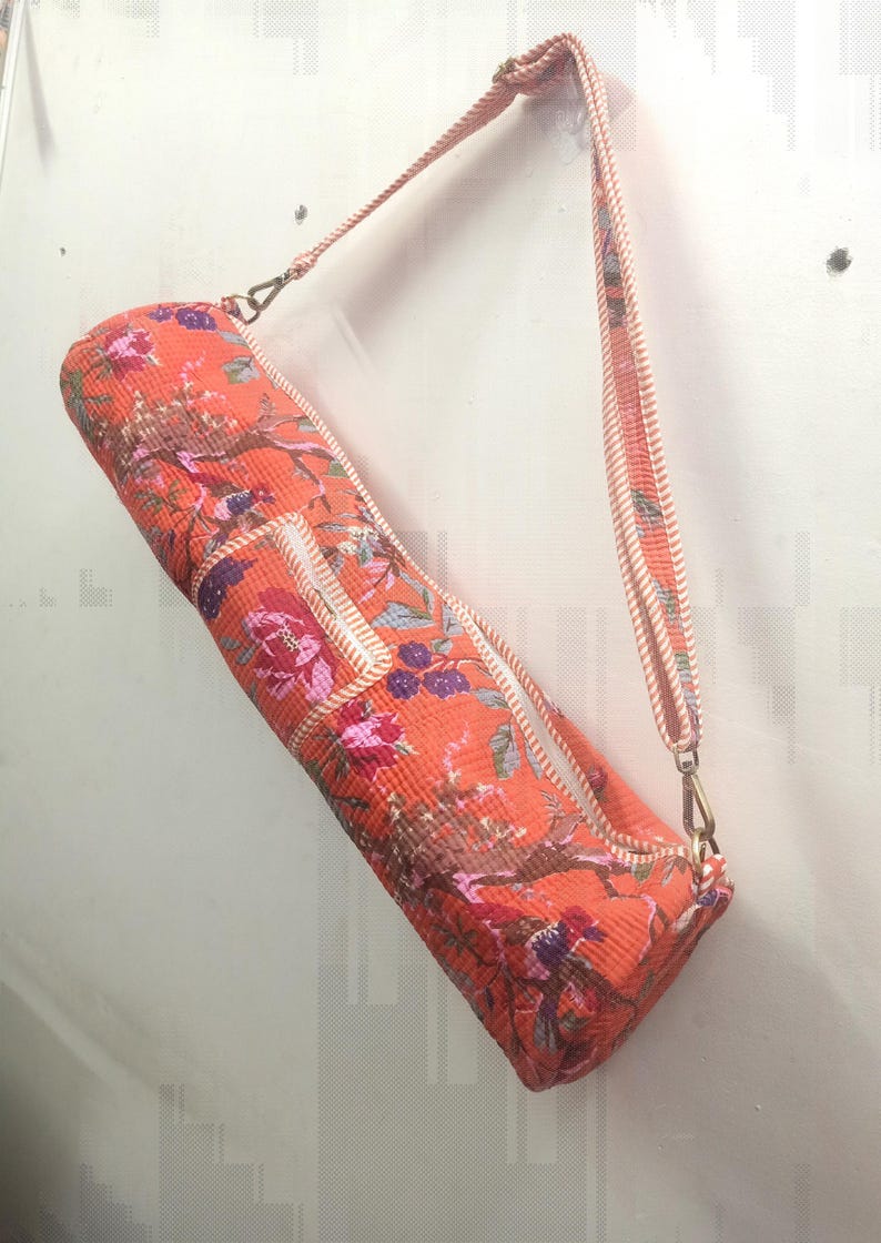 May include: An orange yoga mat bag with a floral pattern in pink, purple, and brown. The bag has a zipper closure and a matching strap with a rope-like design. The bag is hanging against a white wall.