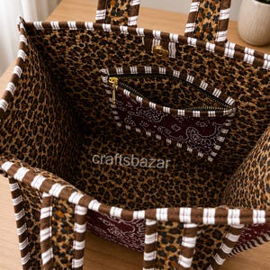 May include: A fabric tote bag featuring a brown and black leopard print interior and exterior, accented with brown and white striped trim on the handles and edges. The bag includes a zipped pocket with a dark red paisley pattern and the text craftsbazar.