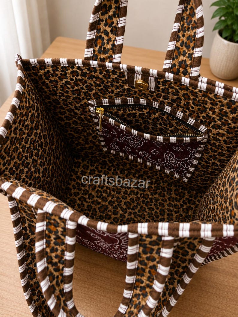May include: A fabric tote bag featuring a brown and black leopard print interior and exterior, accented with brown and white striped trim on the handles and edges. The bag includes a zipped pocket with a dark red paisley pattern and the text craftsbazar.
