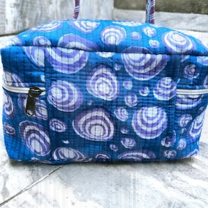 Könnte beinhalten: Eine blaue, gesteppte Kosmetiktasche mit einem sich wiederholenden Muster aus weißen und lila konzentrischen Kreisen. Die Tasche hat einen Reißverschluss und einen Griff mit einem lila-weiß gestreiften Muster. Die Tasche ist rechteckig.