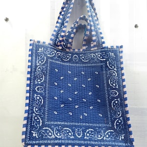 Könnte beinhalten: Eine blaue Tragetasche mit Paisley-Muster und gesteppter Textur. Die Tasche hat ein quadratisches Design mit weißen Paisley-Motiven auf blauem Hintergrund. Die Griffe und Kanten haben ein gestreiftes Muster in Blau und Rosa.